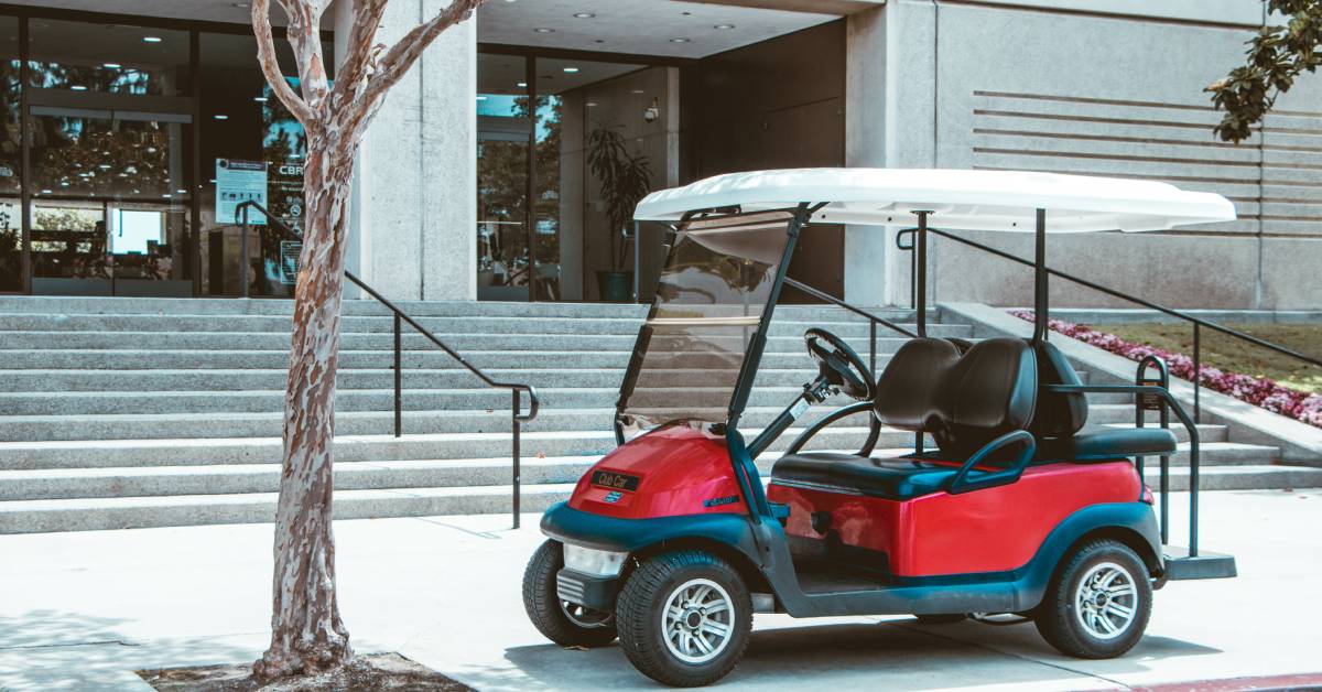 A red street-legal golf cart rental parked outside of a business property with a tree to its left and stairs behind it.