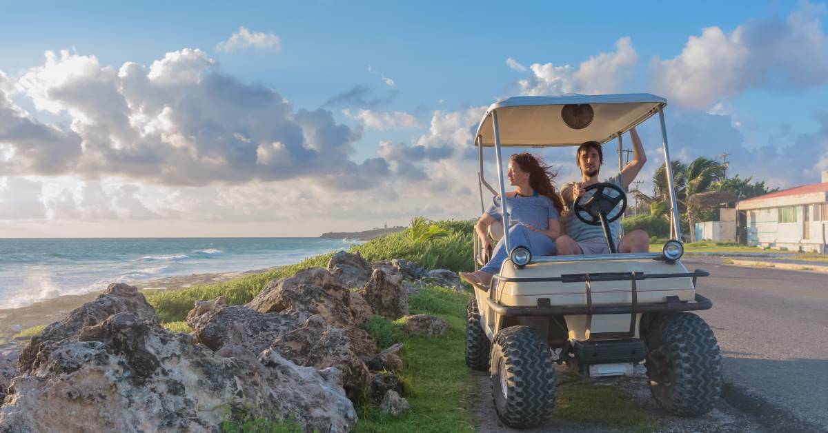Two people sit in a parked street-legal golf cart to watch the sunset over the ocean while clouds pass by.
