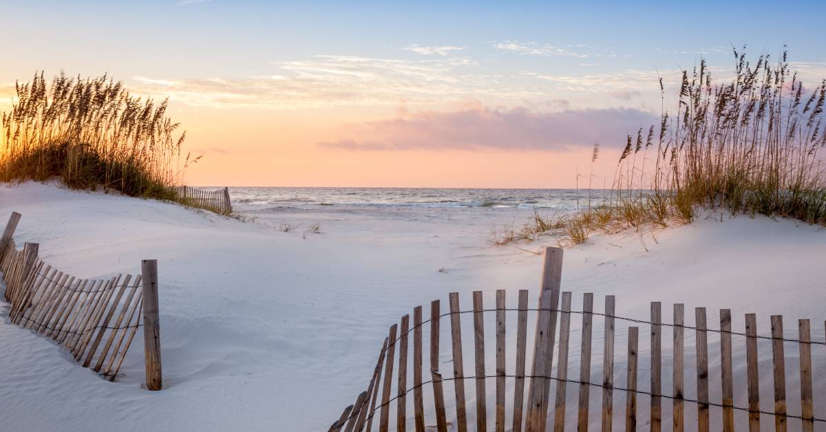 An empty beach with white sand and a small fence. The sky above is turning orange as the sun sets over Florida.