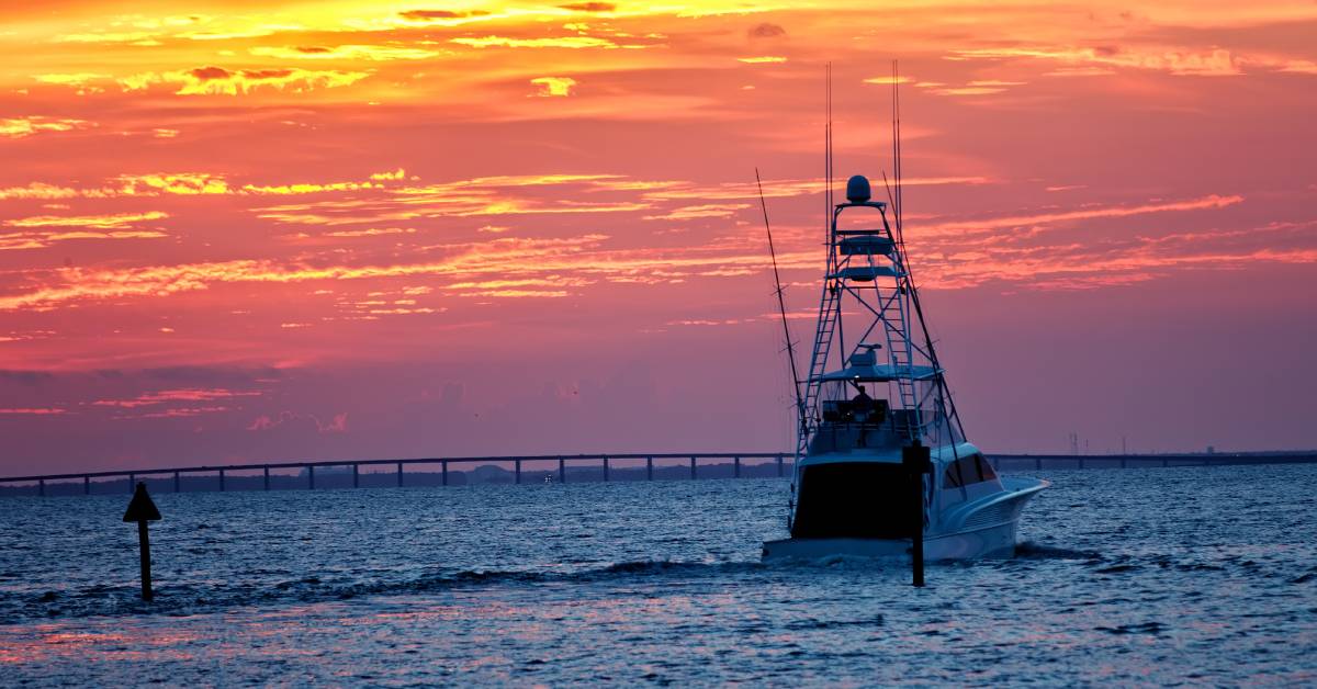 A boat near Destin, FL, navigates away from the shoreline and towards the setting sun while passing a nearby highway.