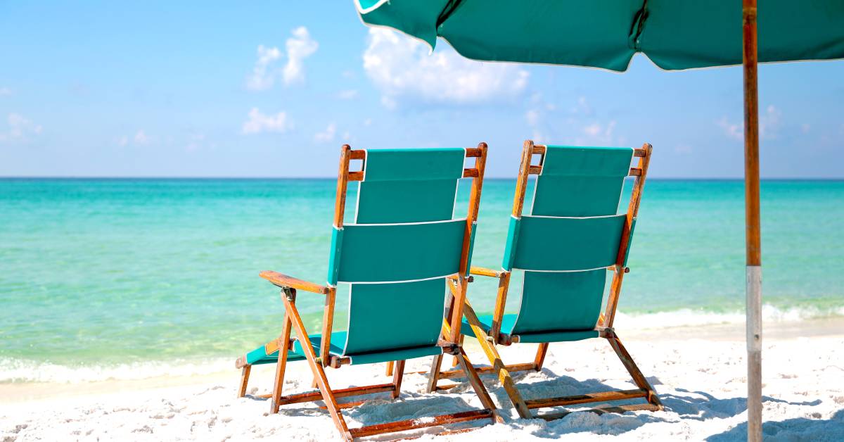 Two identical chairs and one umbrella on a beach in front of the ocean on a sunny day with nobody around.