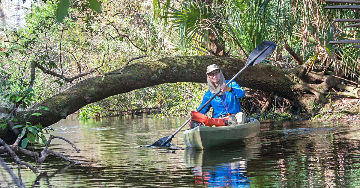 A person wearing a long-sleeved shirt and hat kayaking along a creek in Juniper Lake, FL. There is a branch behind the kayak.