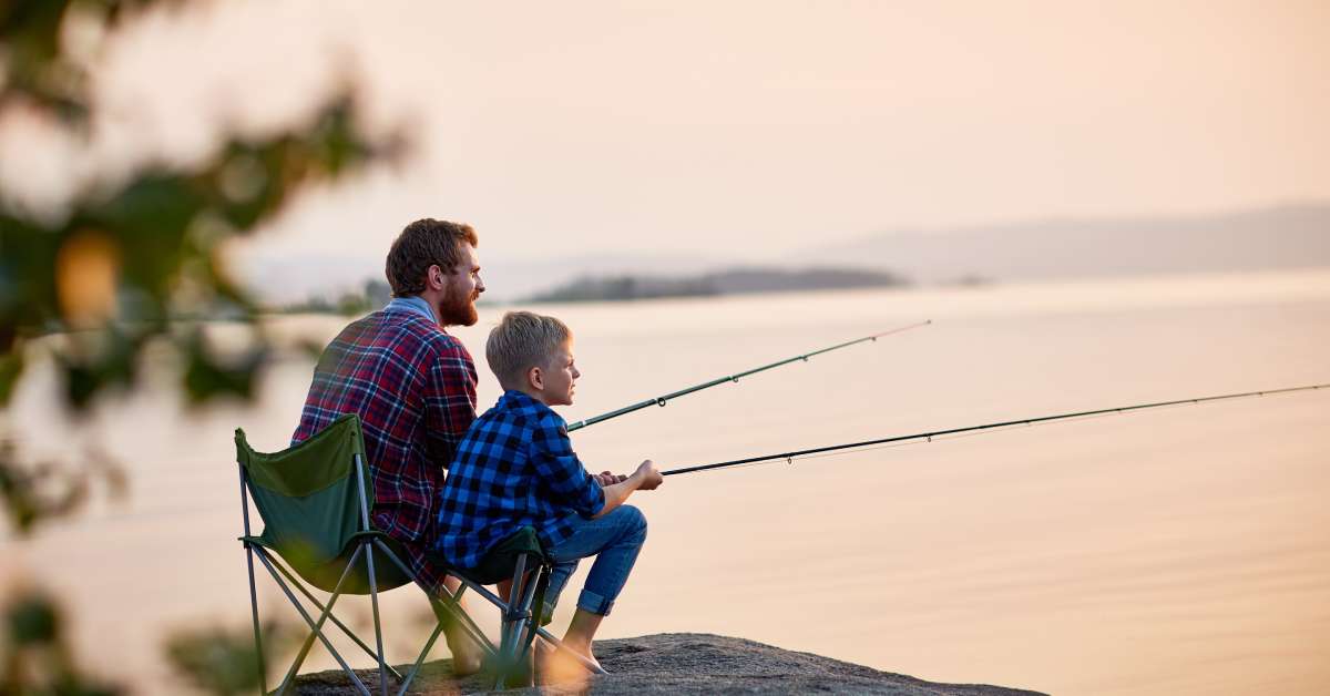 A father and son fishing together from a large rock formation by a lake. The sun is setting over the horizon.