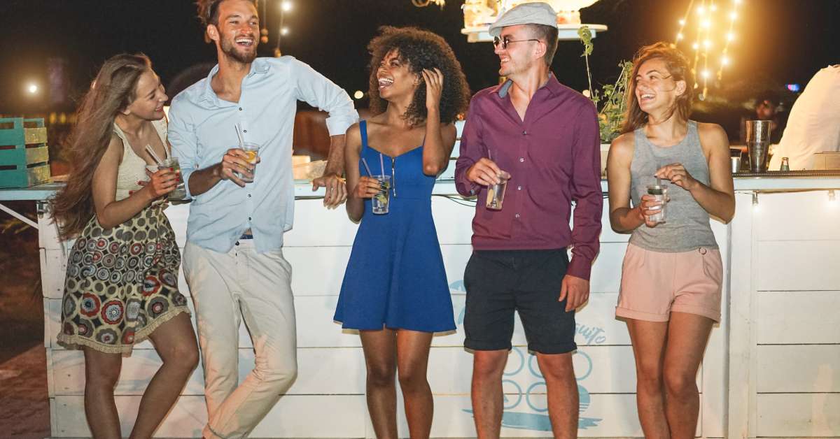 A group of friends stand in front of a white beachside bar while holding drinks in clear cups at night.