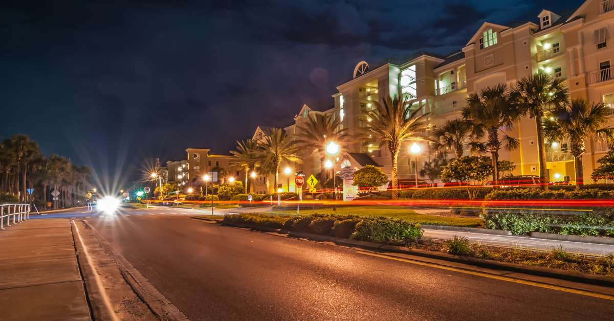 A street at night in Destin, FL, with a car's headlights in the distance. Apartments line the right side of the street.