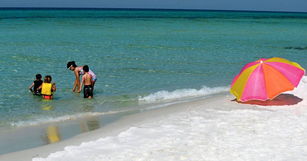 A family plays in the water on a beach during the day. To the right of the family is an umbrella in the sand.