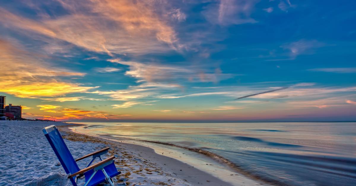 A lounge chair sits on the beach next to the ocean at sunrise. The sky mixes gold and light pink, and the ocean is calm.