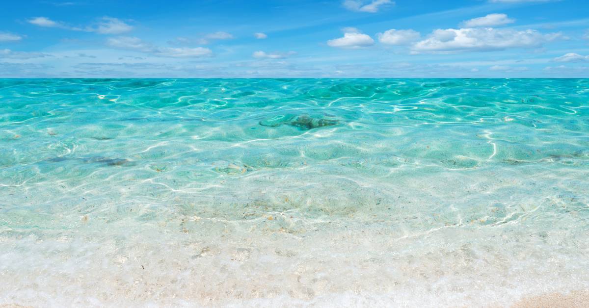 Crystal-clear waters stretch to the horizon beneath a blue sky with clouds. The beachfront sand is light in color.