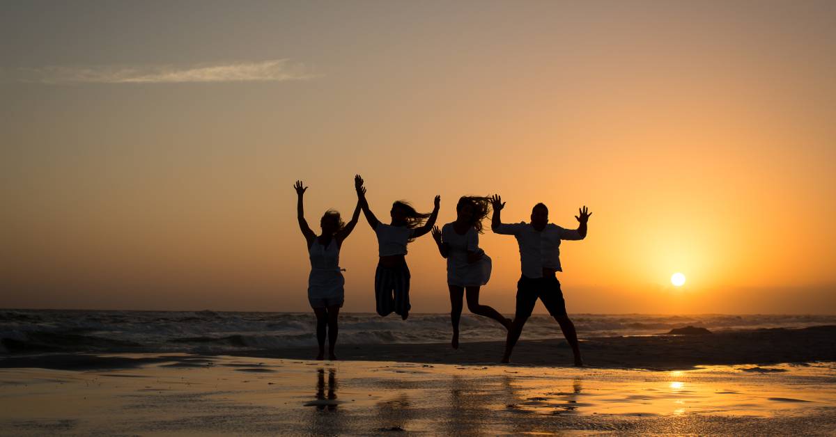 Four people jump together on the shoreline of the beach as the sun sets. Due to the lighting, they're only visible as dark silhouettes.