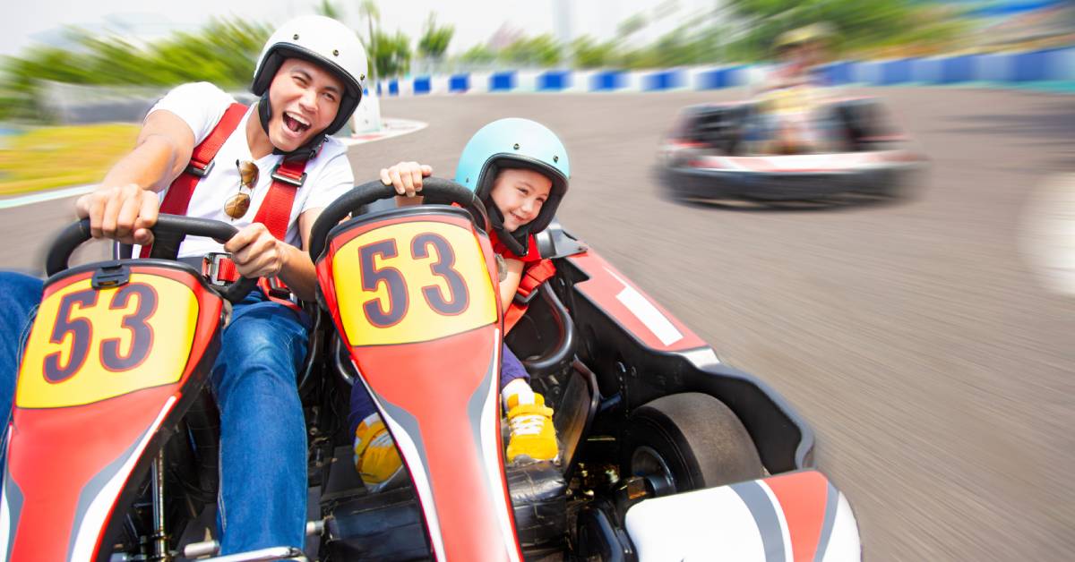 A father and his child drive a go-kart on a fast runway. They wear protective helmets and smile as they steer their car.