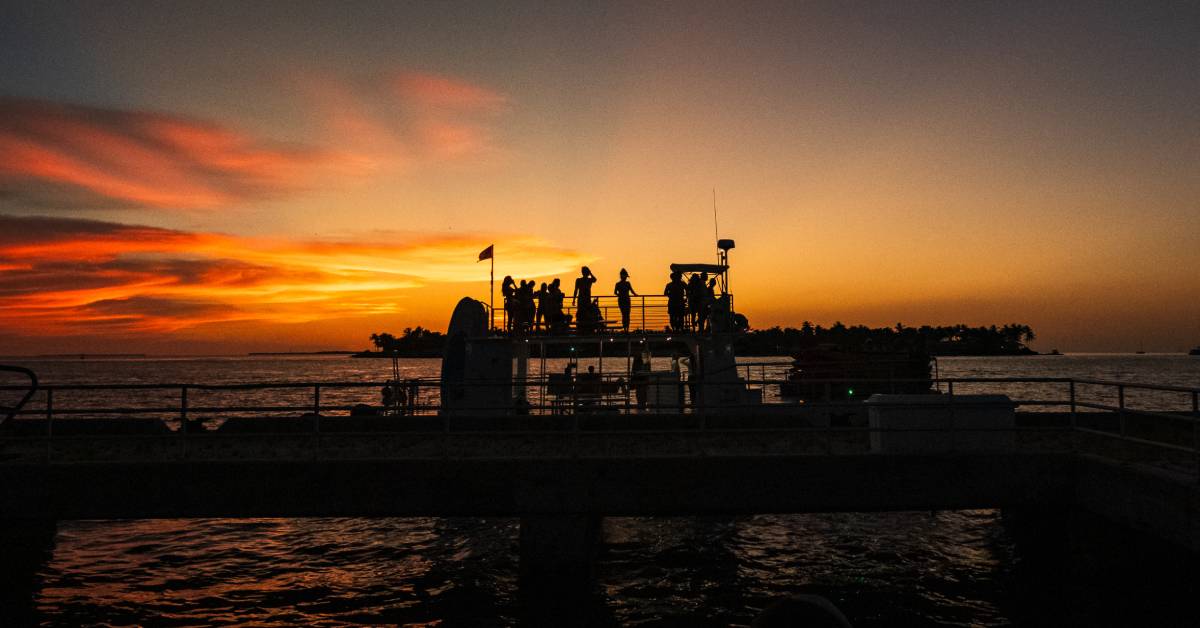 A small cruise boat resting silhouetted in the open ocean as the passengers take in the setting sun on the horizon.