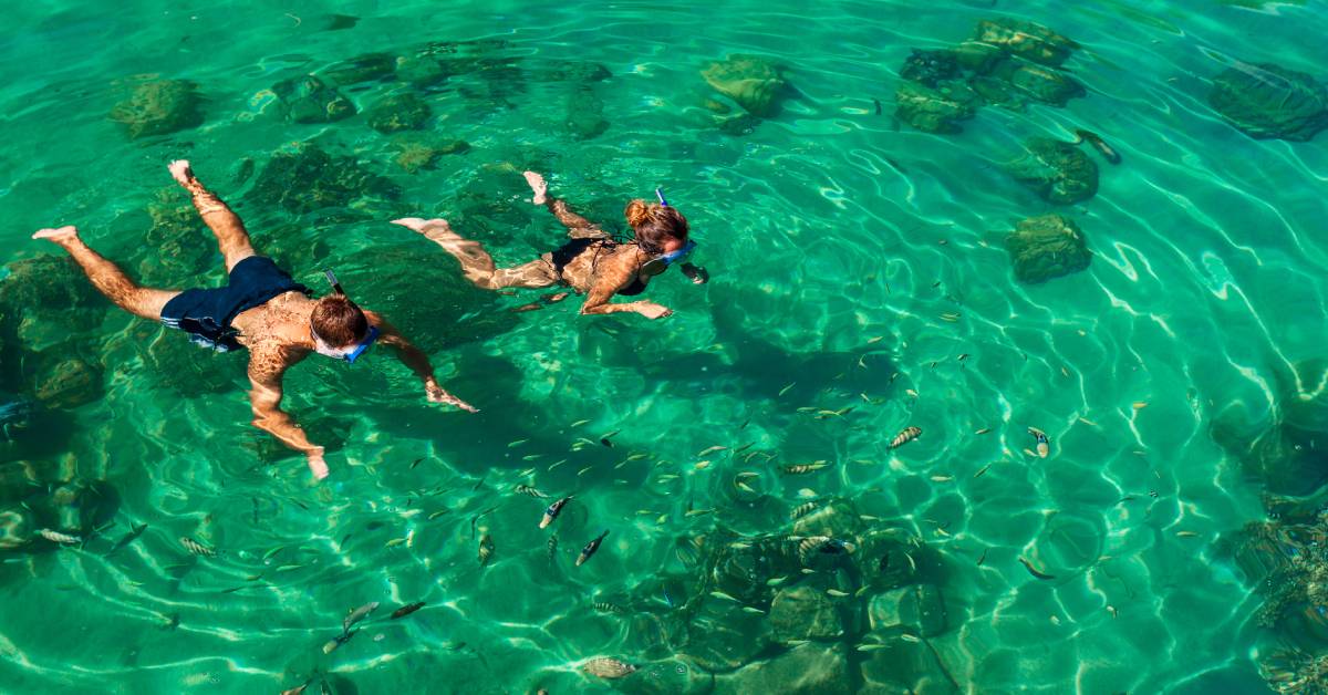 Two young individuals swimming through shallow waters among small fishes and rocks with their snorkeling equipment.