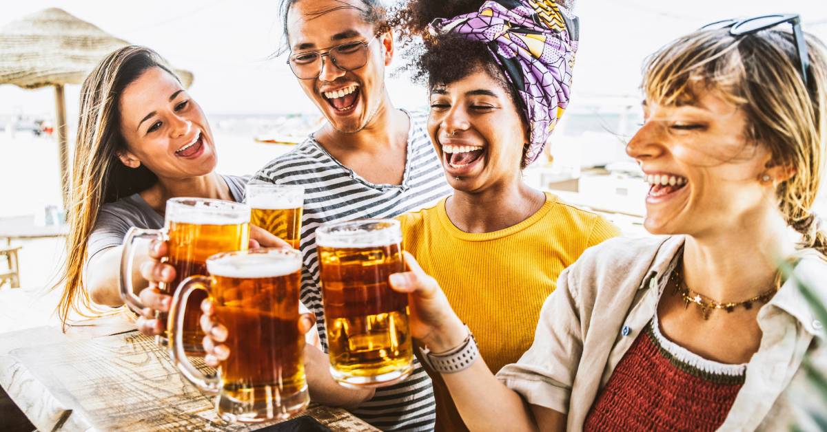 A group of four young tourists toast with their drinks at an outdoor beach bar as they smile and laugh.
