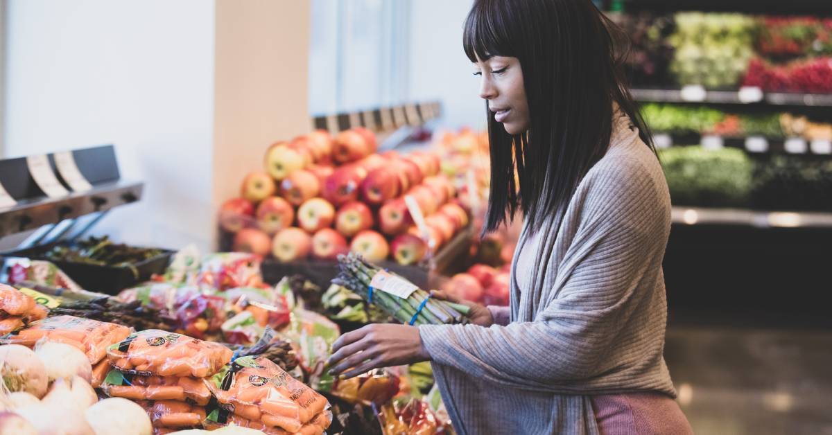 A woman stands over a section of produce as she carefully shops for asparagus and carrots from the store.