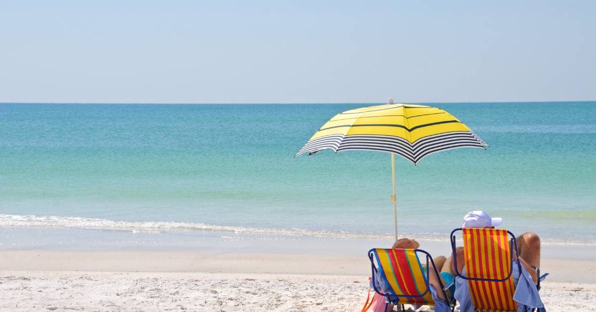 Two people sit on striped beach chairs with a yellow umbrella staked in the sand. They look at the bright blue water.