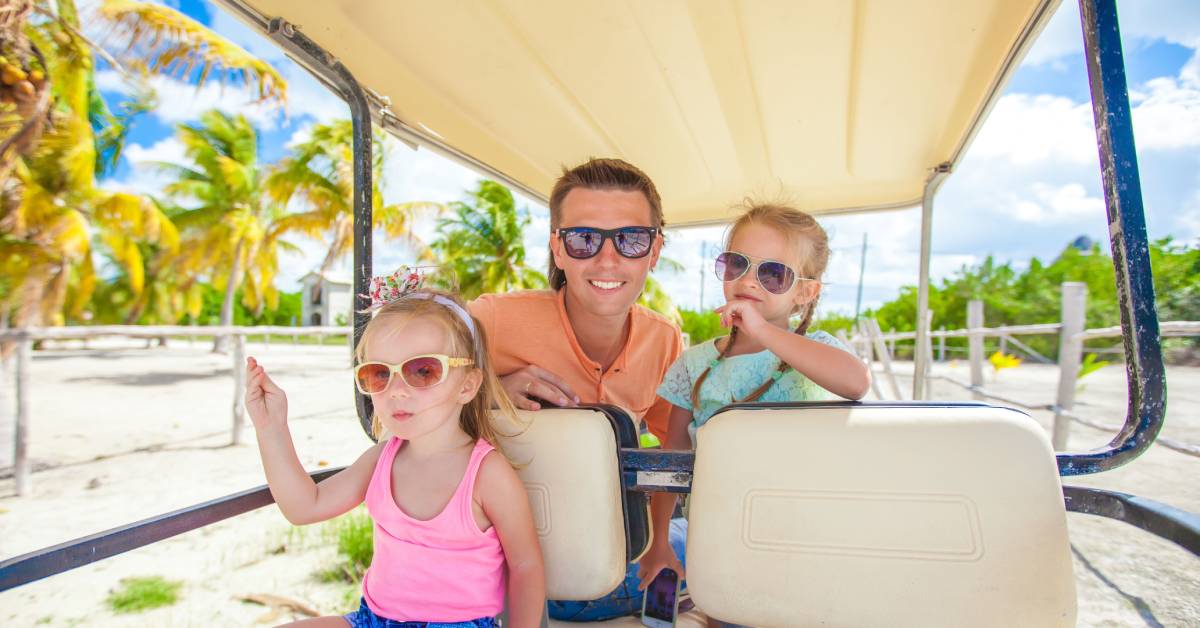 A man and two little girls, all wearing sunglasses, smile as their golf cart shelters them from the sun at a sandy beach.