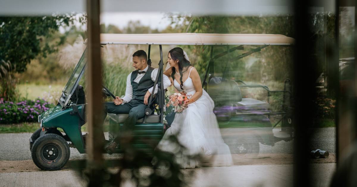 A wedded couple poses as they sit in a golf cart. The bride holds her bouquet, and the groom grabs the steering wheel.