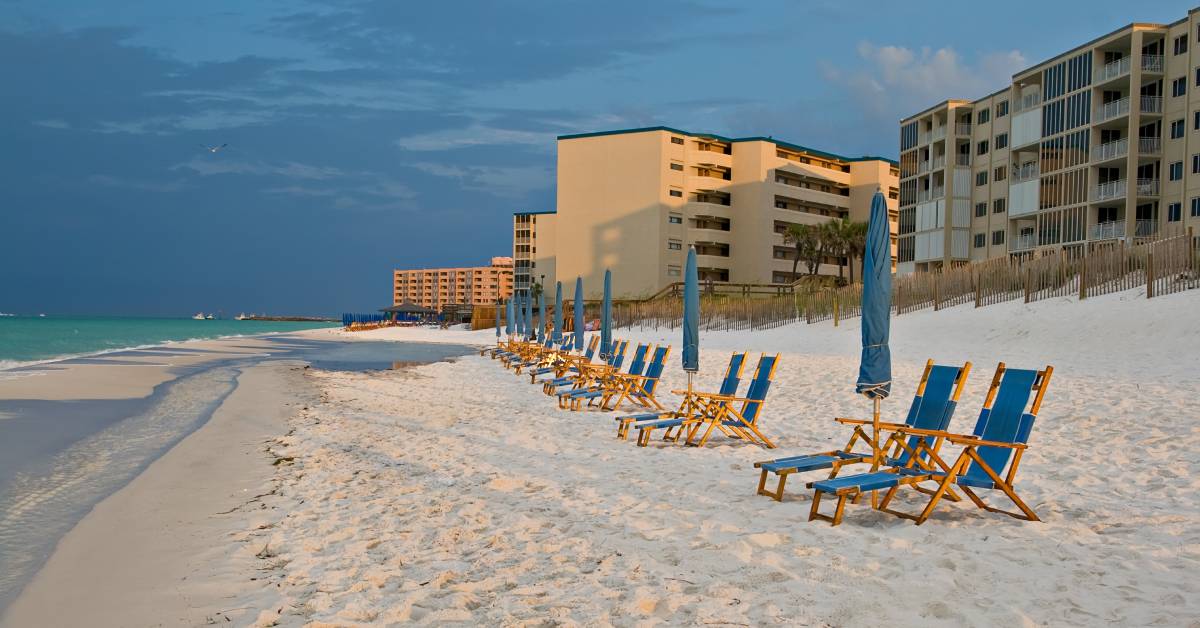An empty sandy beach with lounge chairs lined up and folded umbrellas. There are several buildings in the background.
