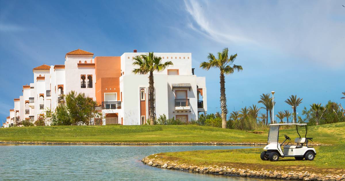 A parked golf cart at the edge of a lake on a sunny day. Trees surround a hotel complex in the distance.