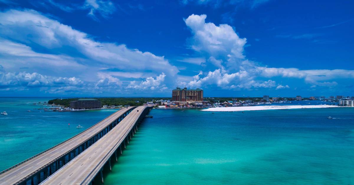 A bridge leading to the town of Destin, Florida. Tan-colored buildings and a cloudy sky are in the distance.