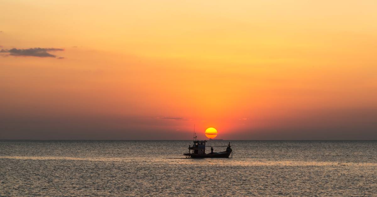 A large fishing boat sitting in the center underneath a sunset. The sky is a deep orange with clouds lining the sun.