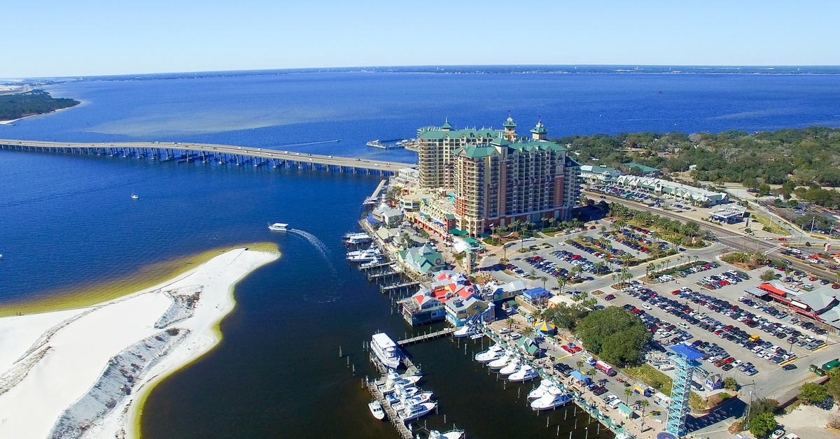 An aerial view of Destin, Florida, on a sunny day with clear skies. A marina, building, peninsula, and ocean are visible.