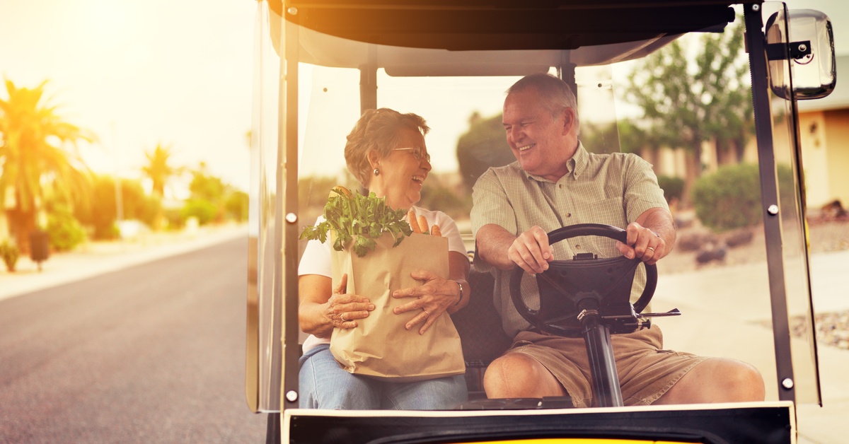 And older couple sitting in a golf cart and smiling at each other as they drive down a main road. The woman is holding groceries.