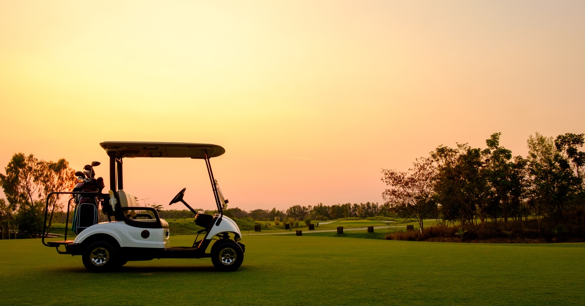 A white golf cart parked on a fairway. The sun has set, and the sky is a blend of pink and yellow.