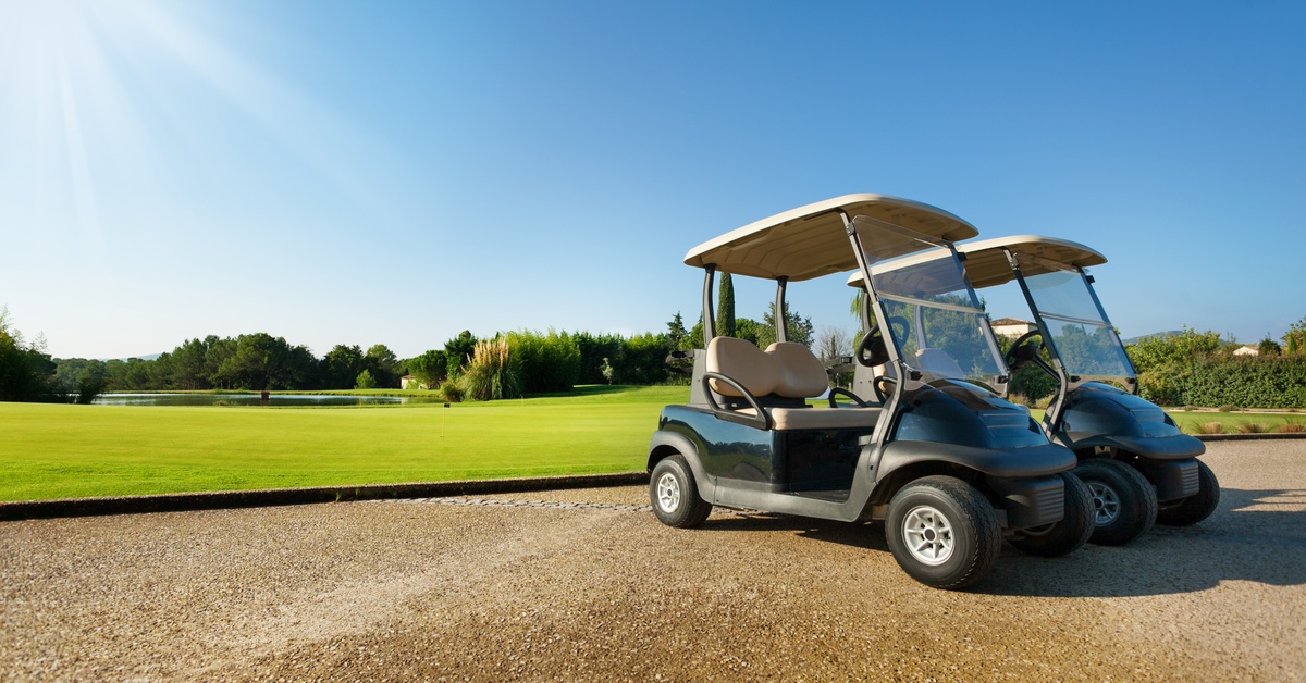 Two green golf carts with sun roofs parked next to each other in front of a golf course on a clear, sunny day.
