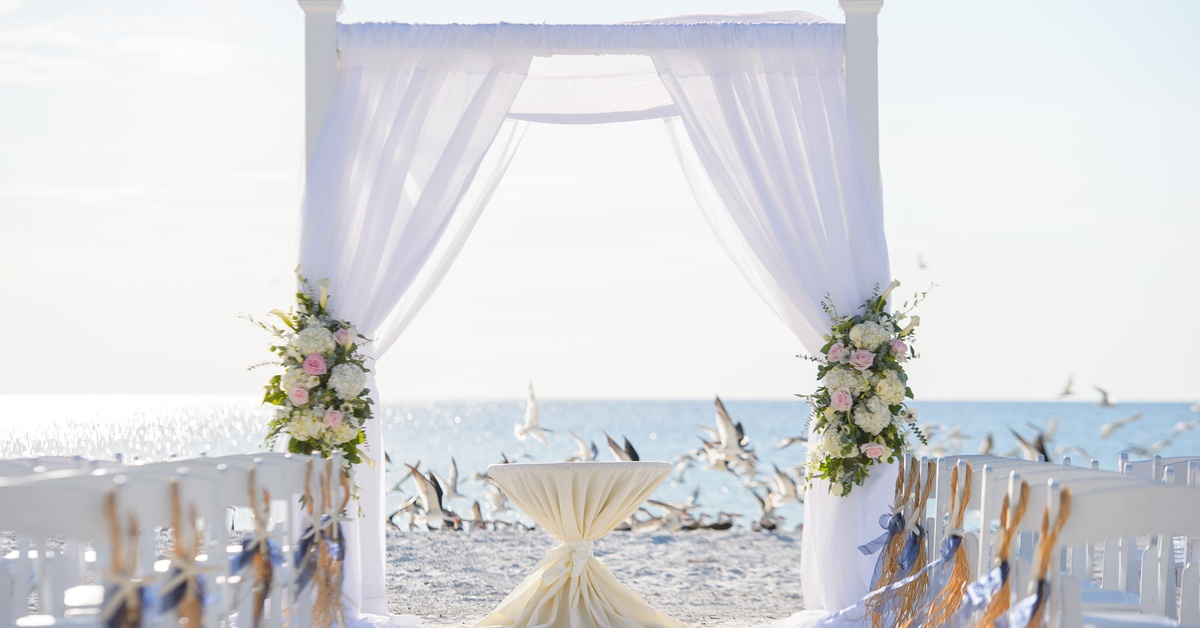 The setup for an ocean-facing beach wedding ceremony with all-white chairs, a pulpit, and a curtained flat arch in broad daylight.