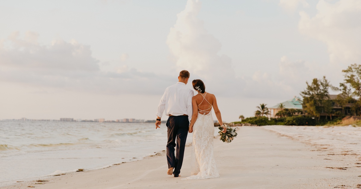 A bride and groom walking hand in hand down the shoreline of a sandy beach with palm trees and buildings in the distance.