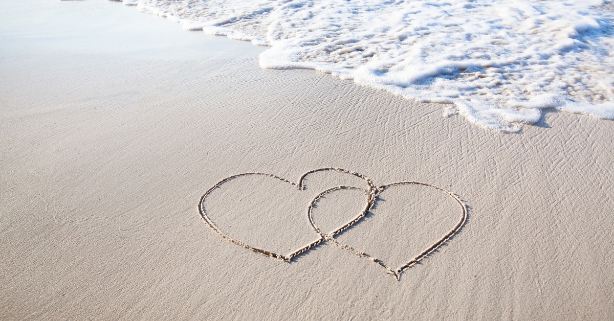 Two hearts drawn into white sand on a shoreline with the froth of the incoming tide above them.